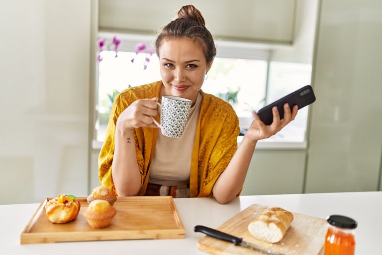 Young beautiful hispanic woman having breakfast drinking coffee using smartphone at the kitchen